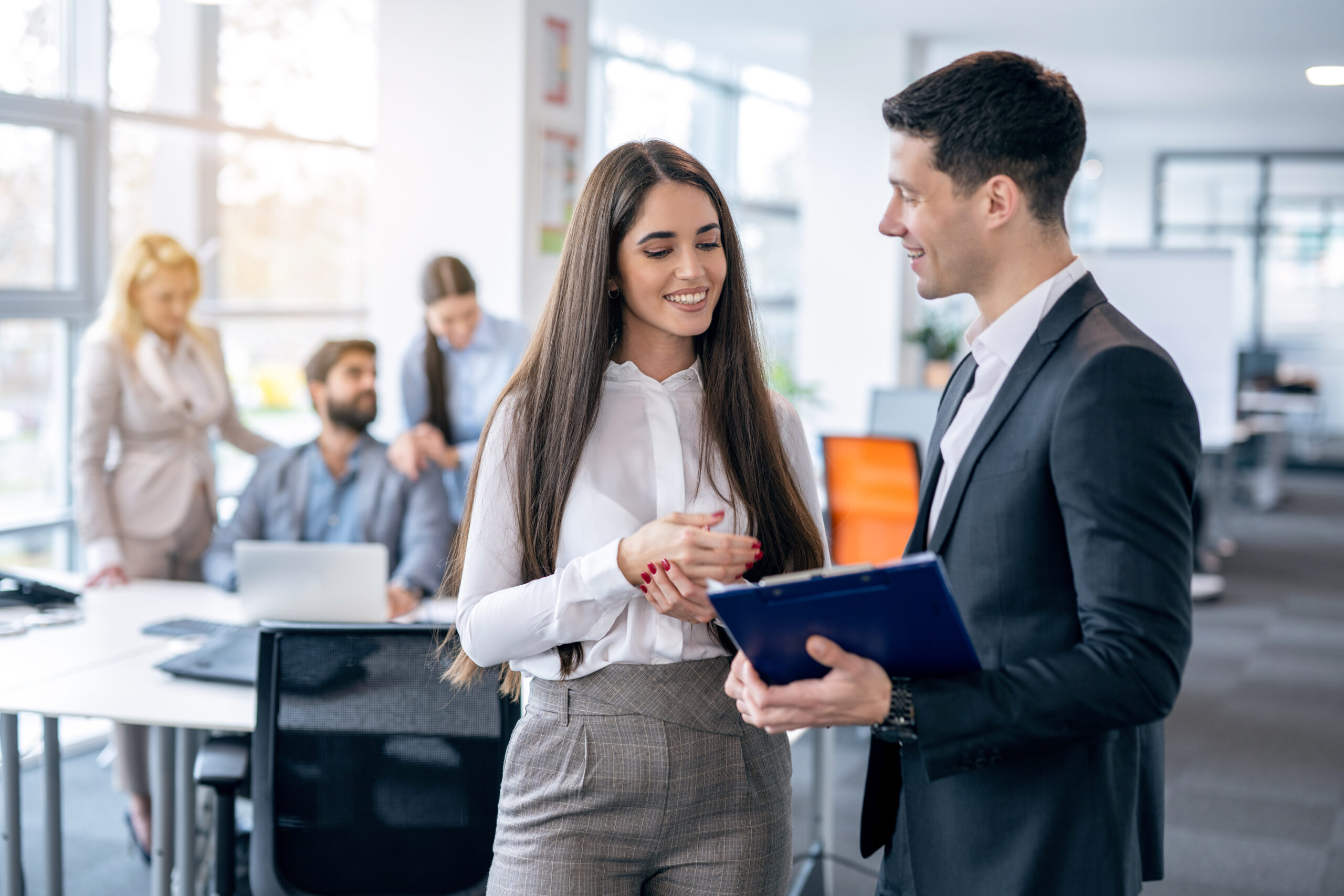 Young business partners talking about company growth, discussing ways to improve business and analyzing data from clipboard during meeting in corporate office with colleagues in the background.